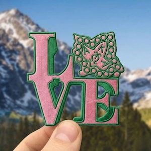 May include: Pink and green embroidered patch spelling "LOVE" with a green emblem. The emblem features Greek letters and a decorative border. The patch is held up against a backdrop of mountains.