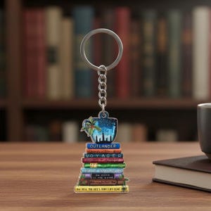 May include: A keychain featuring a stack of books with titles like "Outlander" and "Voyager." The top of the keychain has a blue design with a cityscape and a sprig of holly. The keychain is silver and has a chain link.