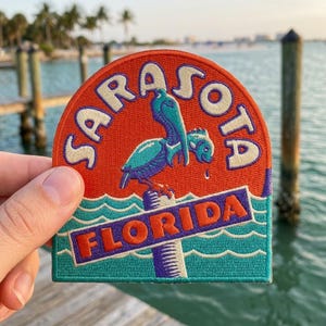 May include: A semi-circular embroidered patch featuring a teal and purple pelican perched on a post against an orange background. The text SARASOTA is arched at the top in white, and FLORIDA is printed in orange on a purple banner over teal waves.