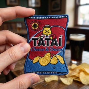 May include: A rectangular embroidered patch shaped like a snack bag with a red and blue background. It features a cartoon character in a hat, the text TÁTAÍ, the words Cheese & Onion, a sunflower icon, and yellow potato chip illustrations.