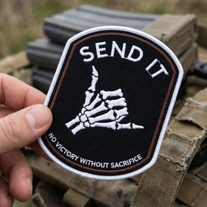 May include: Black patch with white embroidered text "SEND IT" and a skeleton hand giving the shaka sign. The patch has a brown border and the text "NO VICTORY WITHOUT SACRIFICE" in white. The patch is rectangular with rounded corners.