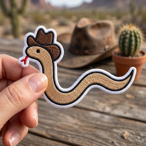 May include: Embroidered patch featuring a cartoon snake wearing a brown cowboy hat. The snake is tan with a black outline and a red tongue. The patch has a white border. A brown cowboy hat and a cactus in a pot are in the background.