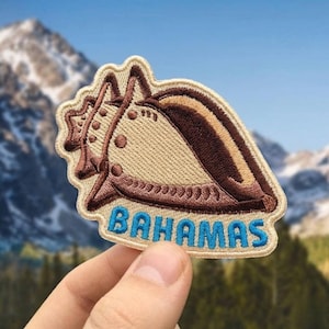 May include: Embroidered patch featuring a conch shell design in tan, brown, and cream colors. The word "BAHAMAS" is embroidered in blue letters at the bottom. The patch is held up against a backdrop of mountains and a blue sky.