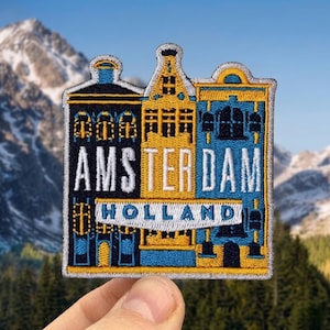 May include: Embroidered patch featuring the word "AMSTERDAM" in yellow and white, with "HOLLAND" below. The patch depicts stylized buildings in blue, yellow, and black, with a mountain and forest background.