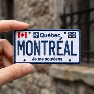 May include: A rectangular embroidered patch designed like a license plate with a white background and dark blue border. It features the text 'MONTRÉAL' in large blue letters, 'Québec' with a fleur-de-lis, a small Canadian flag, and 'Je me souviens'.