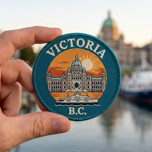 May include: A circular embroidered patch featuring a teal border and the text VICTORIA B.C. The center depicts the British Columbia Parliament Buildings in tan and blue against an orange sky with a sun and a white ferry boat in the foreground.