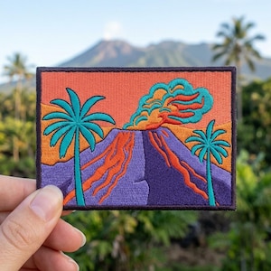 May include: Embroidered patch featuring a volcano erupting with orange lava and teal smoke against an orange and purple sky. Two teal palm trees frame the scene. The patch has a black border.