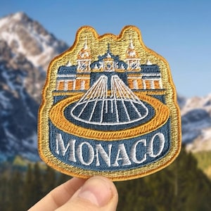 May include: Embroidered patch featuring a detailed illustration of a building with a domed structure and the word "MONACO" in white. The patch has a gold border and a blue and gold design, with a mountain backdrop.