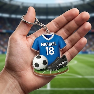 May include: A soccer-themed keychain featuring a blue jersey with "MICHAEL 18", a soccer ball, and a black cleat with "TEAM NAME". The keychain is on a green field background.
