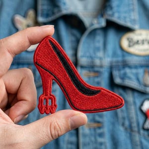 May include: A red embroidered high-heeled shoe patch with a black accent on the side. The patch is held in a person's hand against a denim jacket background. The heel has a unique fork-like design.
