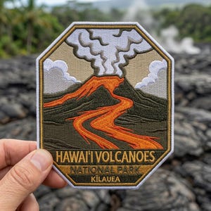 May include: Embroidered patch featuring a volcano erupting with orange lava flowing down a mountain. The patch has the text "HAWAI'I VOLCANOES NATIONAL PARK KILAUEA" in a gold border. The patch is held up in front of a blurred background.