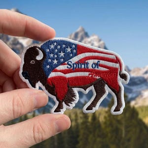 May include: Embroidered patch featuring a bison design with the American flag incorporated into the bison's body. The patch includes the text "Spirit of America" in blue script. The bison is brown and black with a white border.