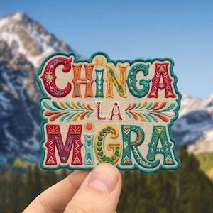 May include: Embroidered patch with the words "CHINGA LA MIGRA" in colorful, stylized lettering. The patch features a cream-colored background with a teal border and decorative floral and sun motifs. The patch is held in a hand against a backdrop of mountains.