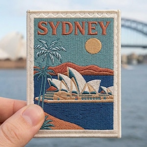 May include: A rectangular embroidered patch featuring the word SYDNEY in orange letters at the top. The design depicts the Sydney Opera House in white and tan, a tall palm tree, a yellow sun, and orange hills against a teal and blue background.