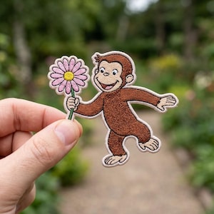May include: An embroidered patch featuring a brown monkey with a smiling expression, holding a pink flower with a yellow center. The patch has a light-colored border and is shaped to follow the outline of the monkey and the flower design.