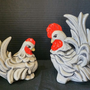 May include: Two ceramic rooster figurines. The roosters are white with red combs and wattles, and yellow beaks. One rooster is sitting, and the other is standing. The figurines have a detailed, textured design.
