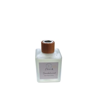 May include: A square glass reed diffuser bottle with a wooden cap and silver ring. The label reads "Clove & Sandalwood" and "200ml Reed Diffuser". The bottle contains a light-coloured liquid.