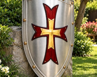 May include: A silver-colored heater shield featuring a central cross design with a deep red background and a gold-toned metallic overlay. The shield is bordered by a twisted metal trim and accented with small, evenly spaced gold-toned circular studs.