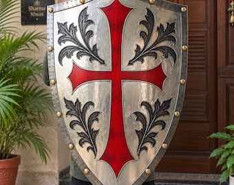 May include: A large, pointed heater shield featuring a metallic silver finish with a prominent red cross centered on the front. The shield is decorated with four black, ornate leaf-like patterns in each quadrant and secured with gold-toned rivets.