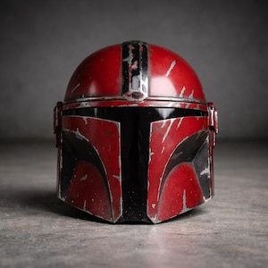 May include: A red and black Mandalorian helmet with a distressed, battle-worn appearance. The helmet features a T-shaped visor and a central vertical stripe. The surface shows scratches and wear, suggesting use. The helmet is a prop or collectible.