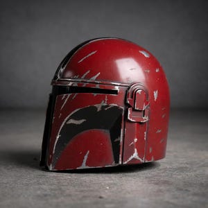 May include: A weathered, red Mandalorian helmet with black accents and silver scratches. The helmet features a T-shaped visor and a rounded top, showcasing a battle-worn aesthetic. The design is reminiscent of a sci-fi space opera.