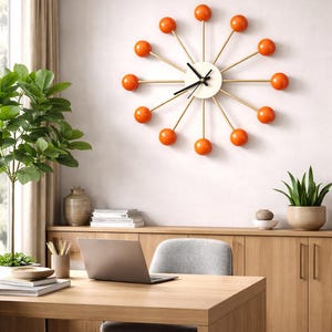 Pode incluir: Um relógio de parede de estilo retro com mostrador branco, ponteiros pretos e raios dourados. O relógio apresenta detalhes esféricos laranja na extremidade de cada raio. O relógio está montado numa parede branca num escritório doméstico.