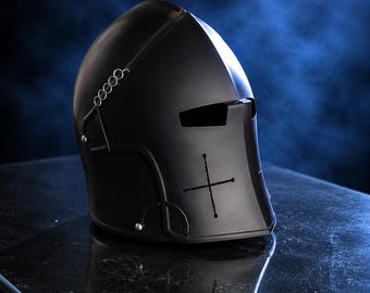 May include: A matte black knight helmet featuring a horizontal eye slit and a vertical cross-shaped ventilation cutout on the front face. The helmet includes metallic rivets and a small chain link detail attached to the side, presenting a smooth finish.