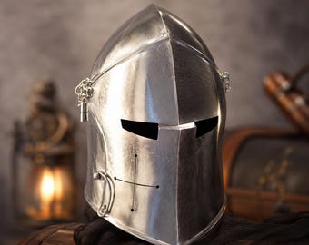 May include: A polished silver-colored metal knight helmet featuring a pointed visor, two horizontal eye slits, and a cross-shaped ventilation pattern on the front. The helmet includes small side chains and a rounded, smooth metallic surface finish.