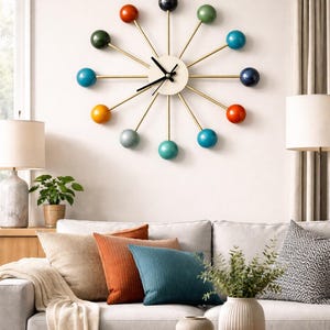 May include: A modern wall clock with a cream-colored face and black hands. The clock features gold-colored arms with colorful spheres in shades of blue, green, orange, and gray. The clock is mounted on a white wall above a gray sofa with colorful pillows.