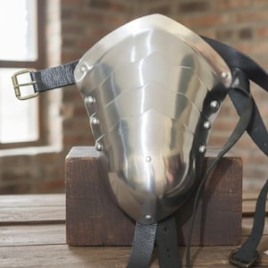 May include: A polished steel shoulder pauldron with riveted plates and a curved design. It features black leather straps with a buckle for attachment. The pauldron is displayed on a wooden block, showcasing its medieval armor aesthetic.