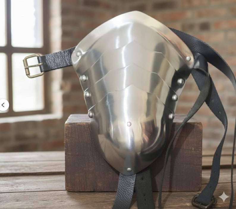May include: A polished steel shoulder pauldron with riveted plates and a curved design. It features black leather straps with a buckle for attachment. The pauldron is displayed on a wooden block, showcasing its medieval armor aesthetic.