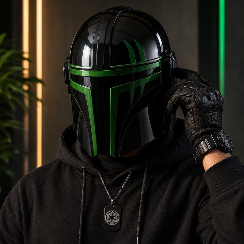 Puede incluir: Un casco negro con detalles verdes, con un acabado brillante. El casco tiene una visera y forma parte de un disfraz. La persona lleva una sudadera con capucha negra y un collar con una placa de identificaci&oacute;n. La persona tambi&eacute;n lleva un guante negro y un reloj.
