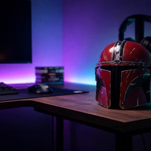 May include: A red and black Mandalorian helmet sits on a wooden desk in front of a computer setup. The helmet has a distressed finish. A black keyboard, mouse, and monitor are visible. Headphones hang in the background, with purple and blue lighting.