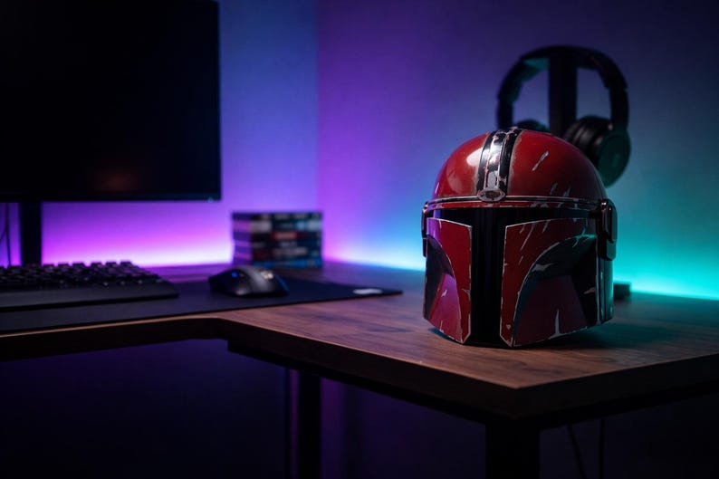 May include: A red and black Mandalorian helmet sits on a wooden desk in front of a computer setup. The helmet has a distressed finish. A black keyboard, mouse, and monitor are visible. Headphones hang in the background, with purple and blue lighting.