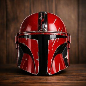 May include: A red and black Mandalorian helmet with silver accents. The helmet has a distressed, battle-worn appearance, with scratches and wear marks visible on the surface. The helmet is set against a dark wood background.