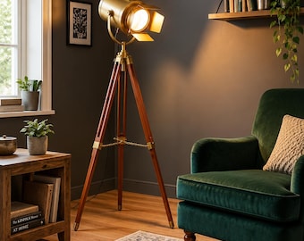 May include: A floor lamp featuring a gold-toned spotlight head with adjustable barn doors, mounted on a dark brown wooden tripod stand with brass hardware accents. The lamp emits a warm light and includes a thin metal chain connecting the three legs.