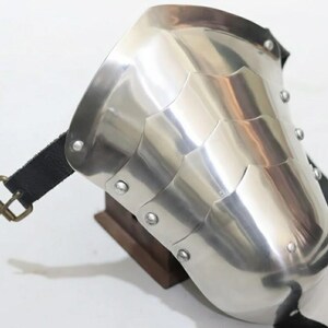 May include: A polished, silver-colored shoulder pauldron with riveted segments. The armor features a curved design and is secured with a black leather strap and a brass buckle. The pauldron is designed to protect the shoulder.