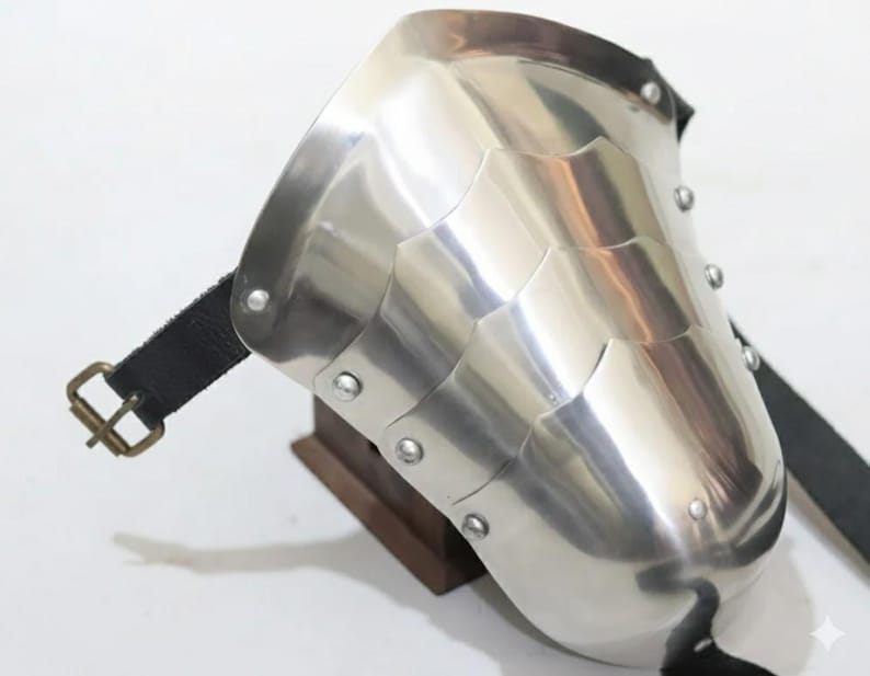 May include: A polished, silver-colored shoulder pauldron with riveted segments. The armor features a curved design and is secured with a black leather strap and a brass buckle. The pauldron is designed to protect the shoulder.
