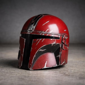 May include: A weathered red and black helmet with silver accents. The helmet has a distinctive design with a visor and a top stripe. The surface shows signs of wear, giving it a battle-worn appearance. The helmet is likely a prop or collectible.