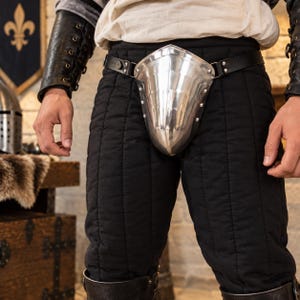 May include: A polished steel codpiece attached to a black leather belt, worn over black quilted trousers. The metal armor piece features a smooth, curved, shield-like design with visible rivets along the edges, secured by a strap around the waist.