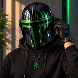 Puede incluir: Un casco negro con detalles verdes, con un acabado brillante. El casco tiene un dise&ntilde;o distintivo con l&iacute;neas angulares y una visera. Se ve un collar con una placa de identificaci&oacute;n negra y un emblema verde. La persona lleva una sudadera con capucha negra y guantes negros.