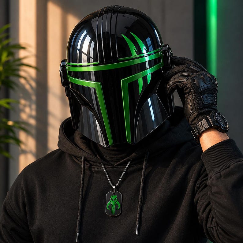 Puede incluir: Un casco negro con detalles verdes, con un acabado brillante. El casco tiene un dise&ntilde;o distintivo con l&iacute;neas angulares y una visera. Se ve un collar con una placa de identificaci&oacute;n negra y un emblema verde. La persona lleva una sudadera con capucha negra y guantes negros.