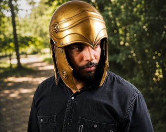 May include: A metallic gold-colored helmet with a smooth, rounded dome, a pointed brow ridge, and side cheek guards. The helmet features circular embossed details on the sides and visible rivets securing the structure, providing full head coverage.