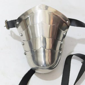 May include: A polished, silver-colored metal arm guard with riveted layered plates. The guard has a curved shape and is secured with a black leather strap and buckle. The design suggests medieval or historical armor.