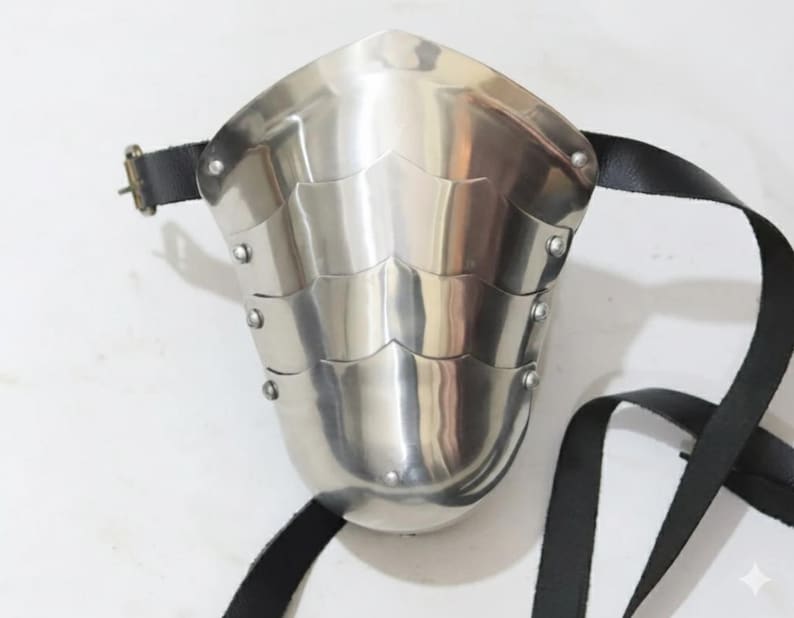 May include: A polished, silver-colored metal arm guard with riveted layered plates. The guard has a curved shape and is secured with a black leather strap and buckle. The design suggests medieval or historical armor.