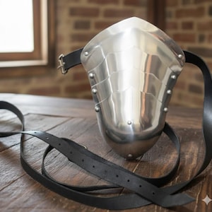 May include: A polished steel pauldron, a piece of medieval armor, with a curved design and riveted segments. It features a black leather strap for attachment. The pauldron is displayed on a wooden surface.