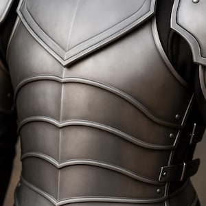 May include: Close-up of a silver-colored chest armor with a layered design. The armor features a V-shaped neckline and horizontal curved plates. Rivets and buckles secure the armor, suggesting a medieval or fantasy theme.