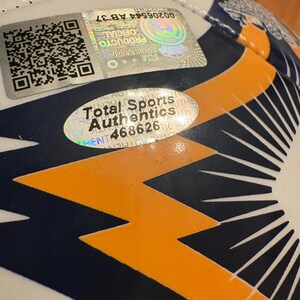 May include: A white soccer ball featuring dark blue and orange geometric patterns. It includes a square Real Madrid holographic verification sticker with a QR code and a separate oval Total Sports Authentics sticker with the number 468626.