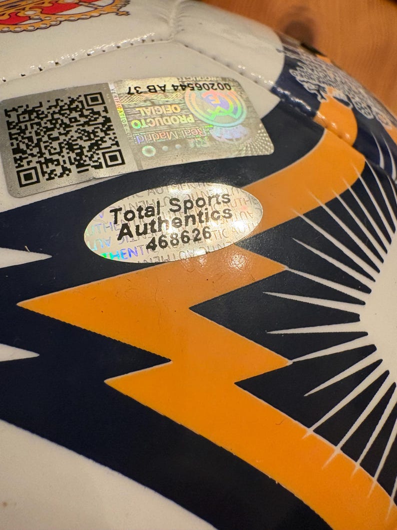 May include: A white soccer ball featuring dark blue and orange geometric patterns. It includes a square Real Madrid holographic verification sticker with a QR code and a separate oval Total Sports Authentics sticker with the number 468626.