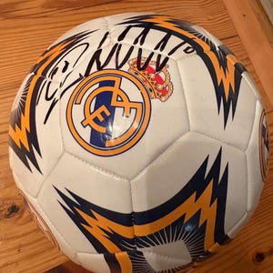 May include: A white soccer ball featuring the Real Madrid club crest in blue, gold, and red, accompanied by a black ink signature. The ball is decorated with dark blue and gold lightning bolt patterns across its multi-paneled surface.